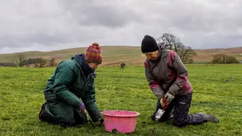 Two women kneeling on a patch of grass holding trowels. There is a pink bucket in front of them, with dirt stains. The women are both wearing waterproof jackets and trousers and woolly hats. They are in an open space with hills in the background. Both are looking to the ground.  