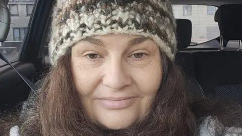 Diane Barnes Diane Barnes is a middle-aged woman. She is sitting in a car and taking a selfie. She has dark hair and is wearing a knitted hat. There are houses in the distance. 