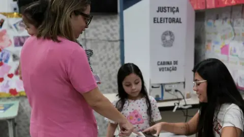 Reuters A voter submits a ballot at a polling station in Brazil as an election worker assists. A voting booth marked “Justiça Eleitoral” stands nearby
