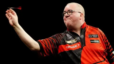 PA Media Stephen Bunting is throwing a dart. A bald man with glasses, he wears a red and black patterned collared shirt adorned with various sponsors' logos.