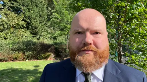 A bald bearded man in an suit stood outdoors