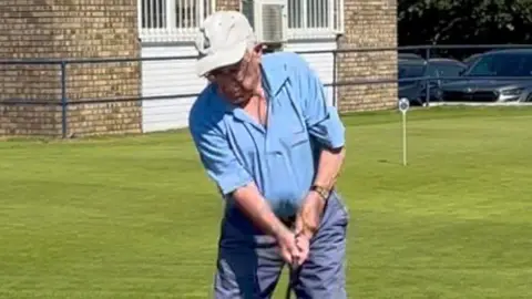 An elderly man getting ready to swing a golf club. He wears a cap, blue polo top and navy blue shorts.
