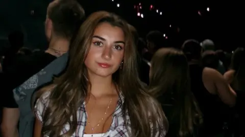 Evie Lewis Evie Lewis, a young woman standing in what appears to be a music venue. She has long dark hair and is wearing a checked short sleeved top over a white vest with a thin gold necklace. Behind her there are lots of people looking at what might be a stage off picture.