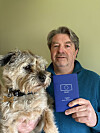 Man holding a blue French passport beside a small shaggy dog indoors.