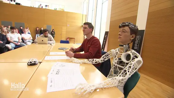Ein Lehrer sitzt neben einem KI-Roboter vor Schülerinnen und Schülern.