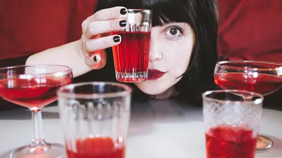 Frau mit schwarz lackierten Nägeln hält sich Glas mit roter Flüssigkeit vor das Auge. Davor Tisch mit rotem Getränk in verschiedenen Gläsern. 