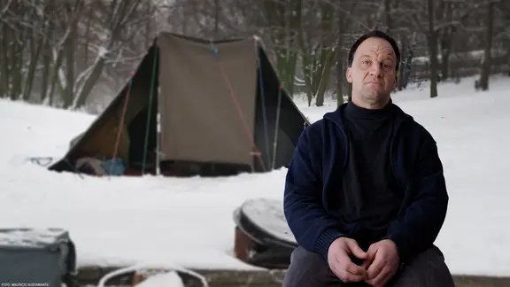 Ein Mann mit Mütze sitzt auf einem Stuhl. Er schaut in die Kamera.