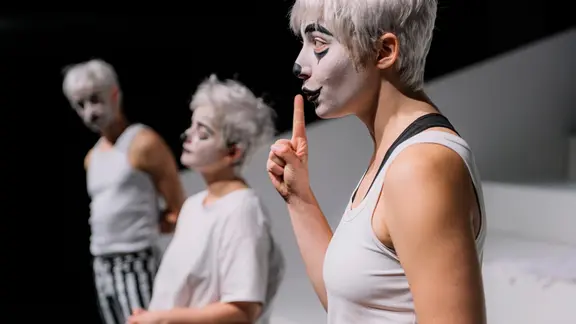 Eine wie ein Clown geschminkte Schauspielerin macht ein Schweigezeichen