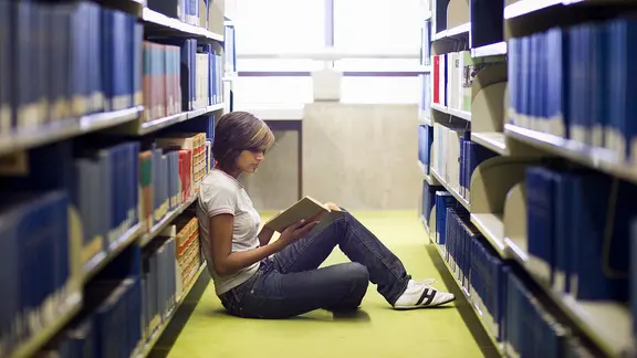 Junge Frau sitzt zwischen den Bücherregalen in der Bibliothek und liest in einem Buch.