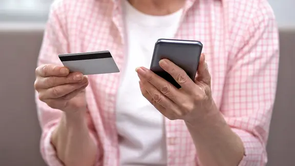 Eine ältere Frau hält eine Kreditkarte in einer und ein Smartphone in der anderen Hand.