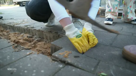 Eine junge Frau hebt einen Pflasterstein aus dem Boden.