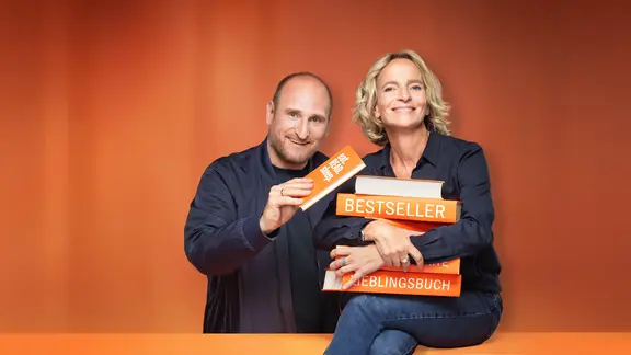 Daniel Kaiser und Katharina Mahrenholtz lächeln mit orangefarbenen Büchern vor orangefarbener Kulisse.
