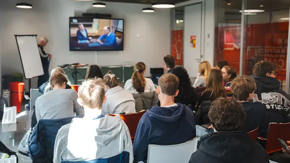 Eine Gruppe von Jugendlichen sitzt in einem modernen Seminarraum und schaut gemeinsam auf einen großen Bildschirm.