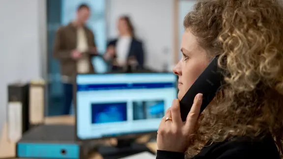 Eine junge Frau telefoniert in einem Büro. Im Hintergrund sind unscharf zwei weitere Mitarbeitende zu sehen.