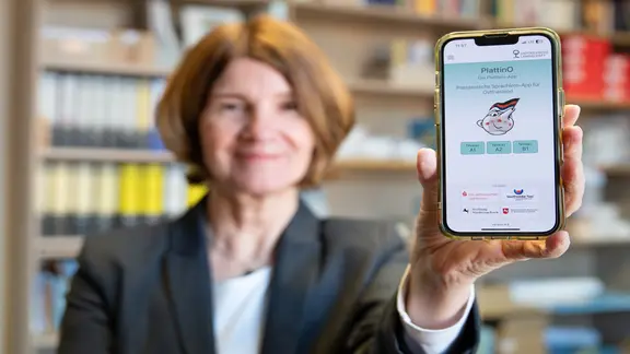 Eine Frau hält ein Smartphone mit ausgestrecktem Arm in eine Kamera, darauf ist die App "PlattinO" zu erkennen.