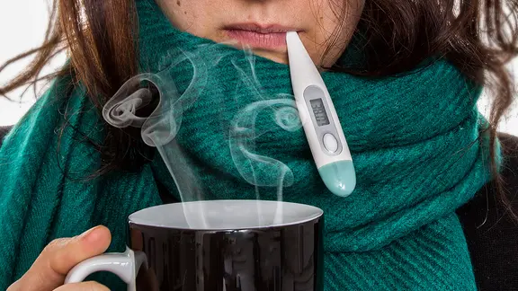 Erkältete Frau mit dampfenden Tee und Thermometer im Mund.