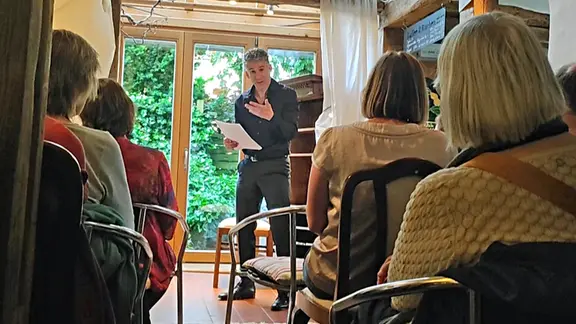 Schauspieler Manuel Klein steht mit einem Blatt Papier in der Hand in einem Raum vor einer Gruppe sitzender Zuschauerinnen. Durch die geöffnete Terrassentür im Hintergrund fällt Tageslicht ins Zimmer.