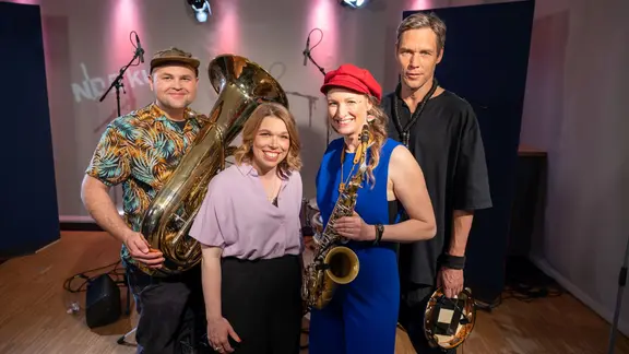 Zwei Männer und zwei Frauen stehen nebeneinander. Ein Mann hält eine Tuba in der Hand und eine Frau ein Saxofon.