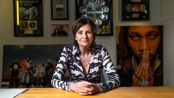 Eine Frau mit braunen halblangen Haaren sitzt mit gefalteten Händen an einem Schreibtisch, im Hintergrund Poster des Wu-Tang Clans. Es ist Eva Ries.