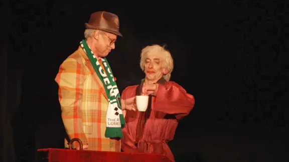 Zwei ältere Männer stehen gemeinsam auf der Bühne, einer mit karierter Jacke, Hut und grünem Schal, der anderem im roten Bademantel