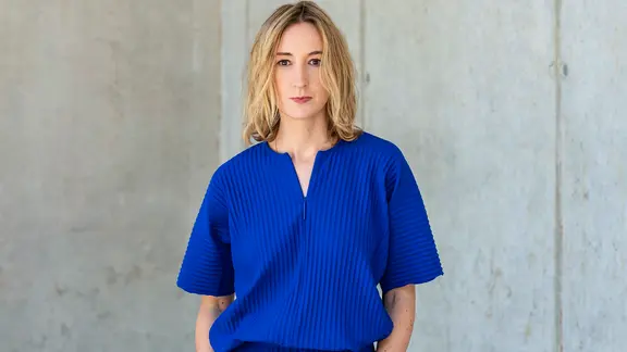 Eine Frau mit blonden Haaren und einer blauen Bluse steht vor einer hellen Wand