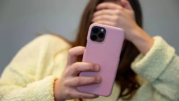 Ein Frau hält sich ein Handy vor das Gesicht und hat gleichzeitig ihre Augen verdeckt.