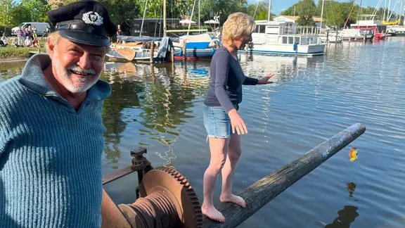 Ein Mann mit Fischerhut lächelt in die Kamera. Neben ihm versucht eine Frau über einen mit Schmierseife eingeriebenen Balken zu laufen.