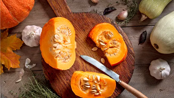 Ein in Stücke geteilter orangefarbener Kürbis mit Messer auf einem Holzbrett
