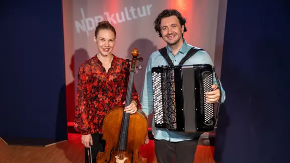 Eine Frau mit rotem Oberteil und einem Cello in der Hand steht neben einem Mann mit einem Akkordeon vor der Brust. Beide lächeln in die Kamera. 