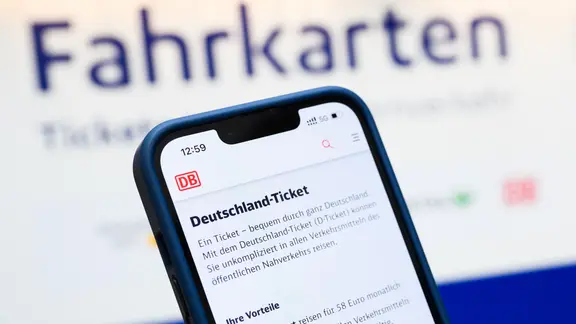 Ein Deutschlandticket wird auf einem Handy angezeigt, im Hintergrund ist ein Fahrkartenautomat.