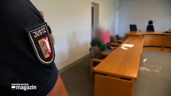 Ein Beamter der Justiz steht in einem Gerichtssaal bei einem Prozessauftakt.