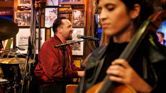 Der Musiker Danger Dan singt und spielt Klavier bei Inas Nacht. Am Bildrand ist eine Cellistin zu sehen, die mit geschlossenen Augen auf ihren Einsatz wartet.