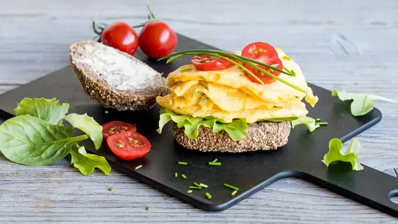 Ein Holzbrett mit Roggenbrötchen, Rührei und Tomaten steht auf dem Tisch.