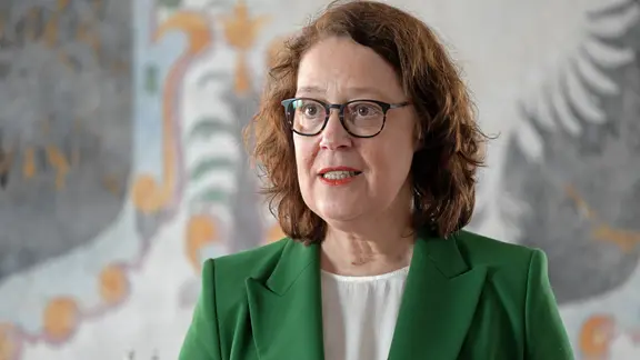 Eine Frau mit grünem Blazer und Brille blickt aufmerksam.