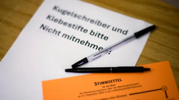 Auf einem Schild steht "Kugelschreiber und Klebestifte nitte nicht mitnehmen", davor Kugelschreiber und ein Stimmzettel