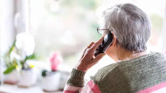 Eine alte Frau steht mit Telefon am Ohr am Fenster.