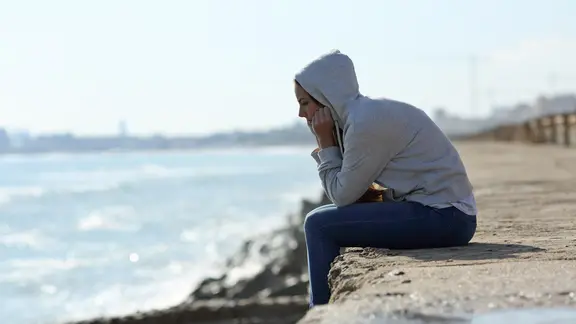 Eine junge Frau mit Kapuzenpullover sitzt nachdenklich auf einer Mole.