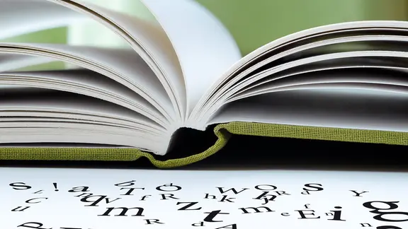 Ein aufgeschlagenes Buch mit weißen Seiten liegt auf einem Tisch, davor liegen Buchstaben.