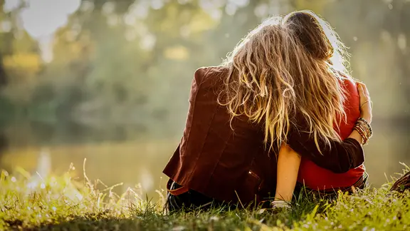 Zwei Frauen sitzen am See, von der die eine ihren Arm um die Schulter der anderen legt.