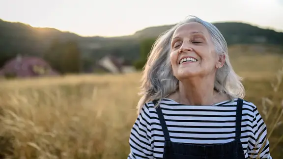 Eine ältere Frau steht vor einem Feld, hat den Kopf in den Nacken gelegt und lächelt mit geschlossenen Augen.