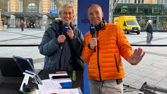 Moderatorin Susanne Stichler und Moderator Stefan Schlag stehen im mobilen Studio am Hauptbahnhof Hannover.