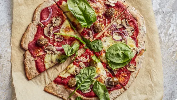 Pizza mit Spinatblättern liegt auf Backpapier auf einer Arbeitsfläche.