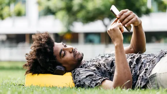 Ein Mann liegt auf einer Wiese, trägt Kopfhörer und hört mit seinem Smartphone Musik.