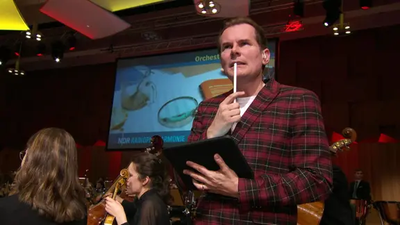 Malte Arkona am Überlegen. Er hält sich einen weißen Stift an die Lippen, vor sich hält er ein Tablet in einer schwarzen Hülle. Hinter ihm sitzt ein Teil eines Orchesters.