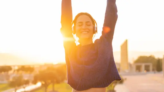 Spanien, Barcelona, Montjuic, glückliche junge Frau, die mit Kopfhörern bei Sonnenuntergang Musik hört.