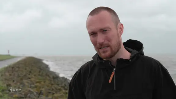 Ein Mann mit sehr kurzen Haaren, Dreitagebart und Windjacke steht an der Nordseeküste.