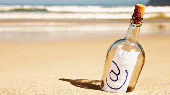 Eine Flaschenpost mit eine Zettel und @-Zeichen steckt halb im Sand an einem Strand.