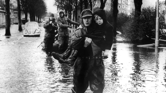 Ein Helfer trägt ein Kind über die überschwemmte Straße nach der Sturmflut 1962