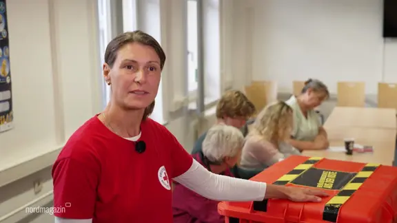 Eine Frau in rotem Shirt mit einem Mikrofon steht neben einer orangefarbenen Kiste mit Warnstreifen und blickt in die Kamera.