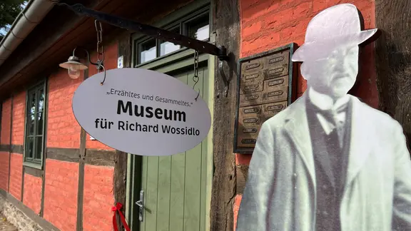 An einem Fachwerkhaus hängt ein Schild, auf dem steht Museum für Richard Wossidlo. Daneben ein lebensgroßes Foto des Forschers.